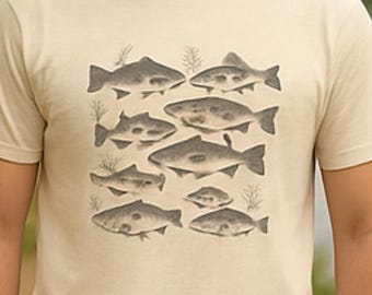 Puede incluir: Camiseta beige de manga corta con un diseño de ilustraciones de peces estilizados en tonos grises. Los peces están dispuestos en un patrón rectangular. La camiseta se lleva al aire libre, cerca de un lago.
