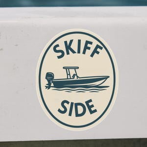 Könnte beinhalten: Ein ovales Aufkleber mit den Worten "SKIFF SIDE" in Dunkelblau, umrandet von einer dunkelblauen Illustration eines Motorboots. Der Aufkleber befindet sich auf einer weißen Oberfläche, mit einem Seil und Bootszubehör.