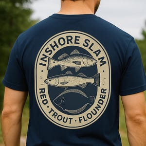 May include: Navy blue t-shirt with a circular graphic featuring three fish and the text "INSHORE SLAM RED TROUT FLOUNDER" in a beige oval. The shirt is short-sleeved and worn by a person wearing a dark blue cap.