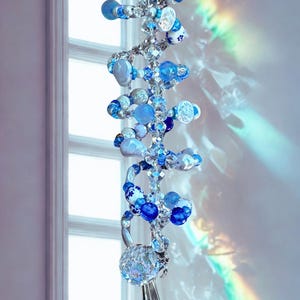 May include: A hanging suncatcher ornament featuring a spiral wire structure adorned with various blue and white beads, including floral patterned ceramic spheres, clear faceted crystals, and smooth glass orbs, ending in a clear crystal prism.