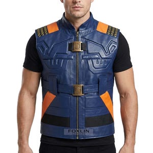 Könnte beinhalten: Eine blaue Lederweste mit orangefarbenen Akzenten und einem Reißverschluss. Die Weste hat ein geometrisches Design, eine schwarze Blende am unteren Rand und goldfarbene Schnallen. Die Schultern haben orangefarbene und schwarze Details. Die Weste wird über einem schwarzen T-Shirt getragen.