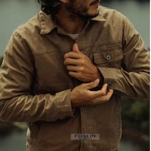 Könnte beinhalten: Eine hellbraune Jacke mit Knopfleiste, zwei Brusttaschen und geknöpften Manschetten. Die Jacke hat einen klassischen Kragen und ist aus strapazierfähigem Material gefertigt. Der Markenname "FOXLIN LEATHERS" ist unten sichtbar.