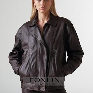 Könnte beinhalten: Dunkelbraune Lederjacke mit Kragen und Reißverschluss. Die Jacke hat ein leicht asymmetrisches Design mit Taschen und einem Knopfverschluss an der Taille. Der Markenname "FOXLIN LEATHERS" ist unten sichtbar.