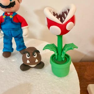 May include: A colorful, handcrafted Super Mario-themed display featuring a Mario figurine in a red cap and blue overalls, a Goomba, and a Piranha Plant in a green pot. The Piranha Plant has a red head with white spots and a white-lined mouth.