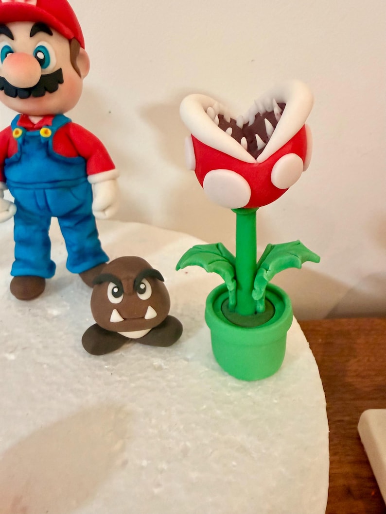 May include: A colorful, handcrafted Super Mario-themed display featuring a Mario figurine in a red cap and blue overalls, a Goomba, and a Piranha Plant in a green pot. The Piranha Plant has a red head with white spots and a white-lined mouth.