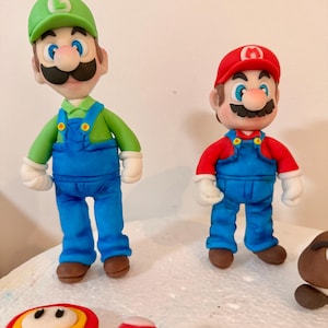 May include: Two figurines of Mario and Luigi, characters from the Super Mario Bros. video game franchise. Mario wears a red hat and shirt with blue overalls. Luigi wears a green hat and shirt with blue overalls. Several mushroom-shaped decorations are in the foreground.