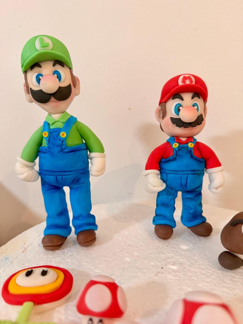 May include: Two figurines of Mario and Luigi, characters from the Super Mario Bros. video game franchise. Mario wears a red hat and shirt with blue overalls. Luigi wears a green hat and shirt with blue overalls. Several mushroom-shaped decorations are in the foreground.