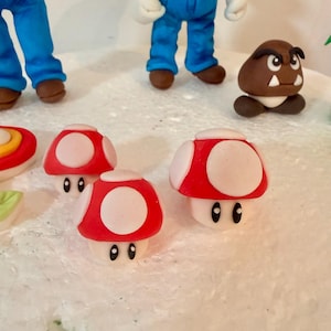 May include: A close-up of a collection of handmade fondant figures, including red and white mushroom decorations, and figures of Mario and a Goomba. The figures are on a white surface.