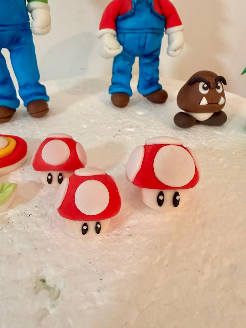 May include: A close-up of a collection of handmade fondant figures, including red and white mushroom decorations, and figures of Mario and a Goomba. The figures are on a white surface.