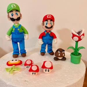 May include: A collection of colorful fondant figures featuring characters from the Super Mario video game series. The figures include Mario, Luigi, a Goomba, a Piranha Plant, and various mushroom designs. The figures are set on a white surface.