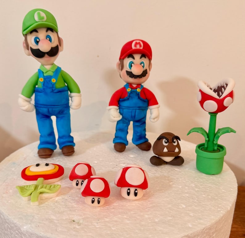May include: A collection of colorful fondant figures featuring characters from the Super Mario video game series. The figures include Mario, Luigi, a Goomba, a Piranha Plant, and various mushroom designs. The figures are set on a white surface.