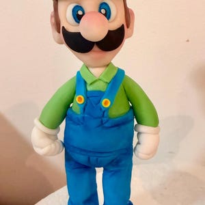 May include: A handcrafted figurine of the video game character Luigi. The figure wears a green cap with a white "L", a green long-sleeve shirt, blue overalls, white gloves, and brown shoes. The character has a black mustache and a pink nose.