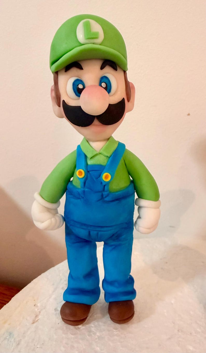 May include: A handcrafted figurine of the video game character Luigi. The figure wears a green cap with a white "L", a green long-sleeve shirt, blue overalls, white gloves, and brown shoes. The character has a black mustache and a pink nose.