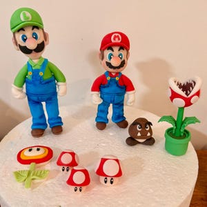 Op de afbeelding: Een set van fondant taartdecoraties met Luigi in een groen shirt en pet, Mario in een rood shirt en pet, een bruine Goomba, een rood-witte Piranha Plant in een groene pot, een vuurbloem en drie rood-witte paddenstoelen met stippen.