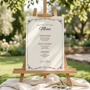 Peut inclure: Un panneau de menu rectangulaire blanc avec du texte noir et une bordure décorative, présenté sur un chevalet en bois clair. Le menu liste les sections Appetizers, Main Course et Dessert, avec les noms Love & Chase imprimés en bas.