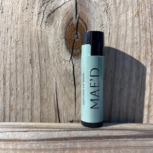 Puede incluir: Un tubo de bálsamo labial natural MAE'D con tapa negra y etiqueta verde claro. El texto "MAE'D" está impreso verticalmente en la etiqueta, junto con "NATURAL LIP BALM". El bálsamo labial está sobre un fondo de madera desgastada.
