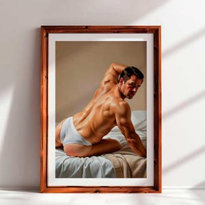 Puede incluir: Una impresión enmarcada que presenta una figura musculosa en calzoncillos blancos, posando sobre una cama con una manta azul claro y beige. La obra de arte está en un marco de madera, apoyada contra una pared blanca.