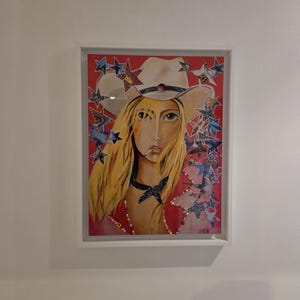 Puede incluir: Obra de arte enmarcada que presenta un retrato de una mujer con sombrero de vaquero. La mujer tiene el pelo largo y rubio y está rodeada de diseños en forma de estrella. El fondo es rojo y la obra está en un marco blanco.
