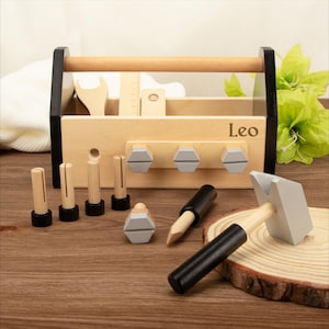May include: A wooden toy toolbox with a black handle and trim, containing toy tools like a wrench, ruler, and hammer. The toolbox is light brown with the name "Leo" printed on it. Additional toy bolts and pegs are included.