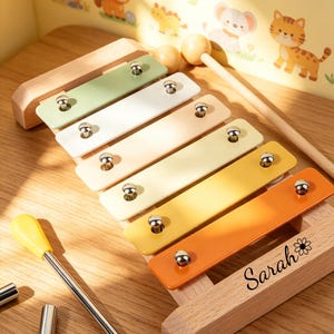 Op de afbeelding: Een houten xylofoon met gekleurde staven in groen, wit, geel en oranje. De xylofoon heeft een houten basis en wordt geleverd met twee hamers. De naam "Sarah" staat op de basis.