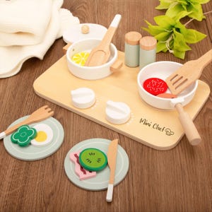 Può includere: Set da cucina giocattolo in legno con tagliere, pentole, padelle, piatti e utensili. Il set include una pentola con una fetta di limone, una padella con cibo rosso e piatti con alimenti. Il tagliere ha il testo "Mini Chef".