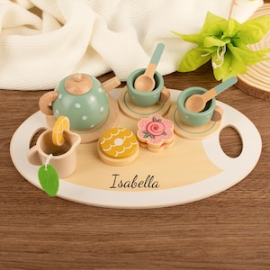 May include: A wooden toy tea set on an oval tray personalized with the name Isabella. The set includes a sage green teapot with white polka dots, two sage green cups with matching saucers, two spoons, a cream pitcher with a lemon slice, and two cookies.