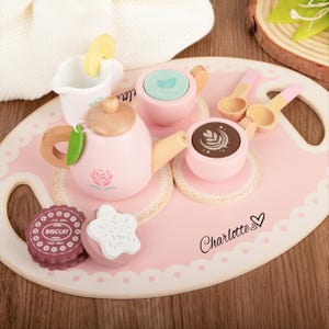 Könnte beinhalten: Ein hölzernes Teeservice mit einem rosa Tablett, einer Teekanne, einer Tasse und Untertassen. Die Teekanne hat ein Rosenmuster, und das Set enthält Holzlöffel und einen Keks. Auf dem Tablett steht der Name "Charlotte".