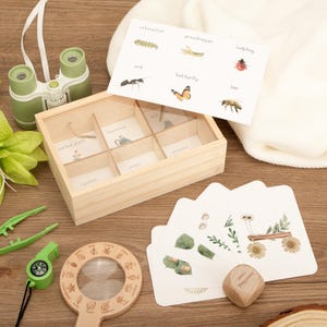 May include: A wooden educational toy set with a magnifying glass, binoculars, and a wooden box with compartments. The box contains cards with insect illustrations and labels. Additional cards with botanical illustrations are also present.