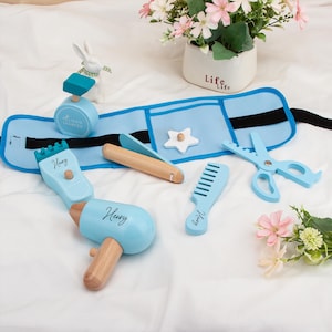 Pode incluir: Um conjunto de brinquedos de barbeiro de madeira azul claro com um avental azul a condizer. O conjunto inclui um secador de cabelo, um pente, tesouras e outros acessórios. A palavra "Henry" está escrita no secador de cabelo e no pente. Um coelho de cerâmica branco e um vaso de flores estão no fundo.