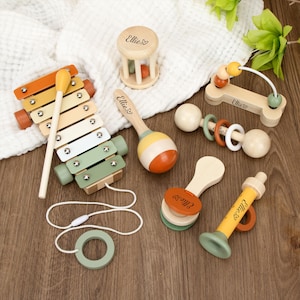 May include: A collection of wooden baby toys, including a xylophone, maracas, and a rattle. The toys are painted in pastel colors of orange, yellow, green, and white. Each toy is engraved with the name "Ellie".