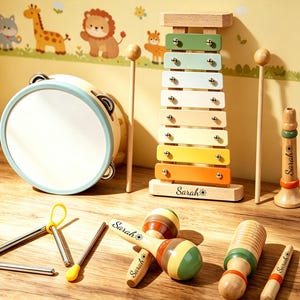 Op de afbeelding: Een verzameling houten muziekinstrumenten, waaronder een tamboerijn, een xylofoon, maracas en een fluit. De xylofoon heeft kleurrijke toetsen en de naam "Sarah" is op de basis gegraveerd. De instrumenten zijn op een houten oppervlak geplaatst.