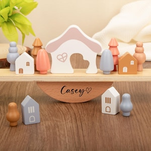 May include: A pastel-coloured wooden toy set, including a house, trees, and other shapes. The set features a wooden balance board with the name "Casey" and a heart symbol. The toys are arranged on a wooden surface.