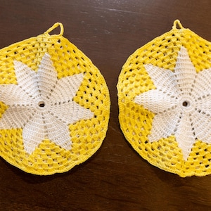 Può includere: Due presine fatte a mano all'uncinetto giallo e bianco. Ogni presina presenta un motivo a stella bianca al centro, circondato da un tessuto aperto giallo. Un piccolo anello è in alto per appenderle. Le presine hanno un diametro di circa 18 cm.