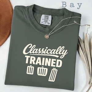 Könnte beinhalten: Ein gefaltetes, olivgrünes T-Shirt mit dem weißen Aufdruck "Classically TRAINED" und Pedal-Grafiken. Eine silberne Halskette mit Anhänger liegt auf dem Shirt. Das Wort "Bay" steht oben.