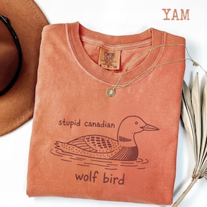 May include: A coral-colored t-shirt with the words "stupid canadian wolf bird" and a graphic of a loon. The shirt has a crew neck and a small tag. A gold necklace with a pendant is draped across the shirt.