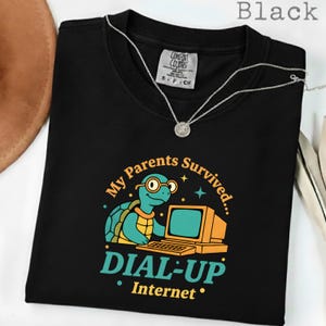 Könnte beinhalten: Schwarzes T-Shirt mit einem Cartoon-Schildkröten-Design mit Brille und Computer. Der Text lautet: "My Parents Survived... DIAL-UP Internet." Eine silberne Halskette liegt auf dem Shirt.