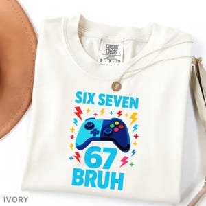 Könnte beinhalten: Elfenbeinfarbenes T-Shirt mit einem blau-marinefarbenen Videospiel-Controller-Motiv. Der Text "SIX SEVEN 67 BRUH" ist blau. Eine goldene Halskette und ein brauner Hut sind sichtbar. Marke Comfort Colors.