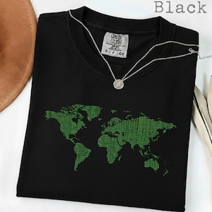 May include: Black t-shirt with a green world map design made of binary code. The shirt has a crew neck and short sleeves. A silver necklace with a round pendant is draped over the shirt. The word "Black" is printed at the top.