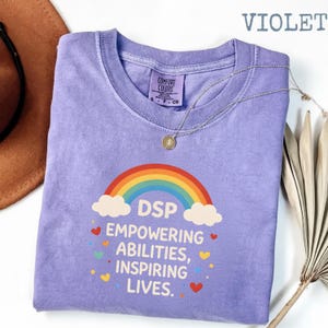 May include: A violet Comfort Colors t-shirt featuring a rainbow graphic and the text "DSP Empowering Abilities, Inspiring Lives." A silver necklace with a pendant is draped over the shirt.