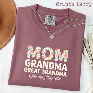 May include: A mauve-colored Comfort Colors t-shirt with the words "MOM GRANDMA GREAT GRANDMA I just keep getting better" in white and floral print. A silver necklace with a pendant is draped on the shirt.