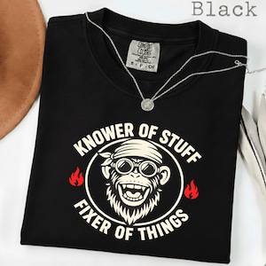 May include: Black crew neck t-shirt with a graphic of a monkey wearing sunglasses and a bandana, with the text "KNOWER OF STUFF FIXER OF THINGS" in a white circular design. A silver necklace is draped across the shirt.