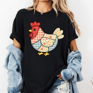 Könnte beinhalten: Schwarzes T-Shirt mit einem bunten Patchwork-Hühnerdesign. Das Huhn hat einen roten Kamm, einen gelben Schnabel und ist mit Blumen- und Vichy-Mustern in Rot, Blau und Gelb verziert. Das T-Shirt hat kurze Ärmel.