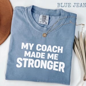 May include: A blue jean coloured Comfort Colors t-shirt with the text "MY COACH MADE ME STRONGER" in white. A silver necklace with a circular pendant is draped across the shirt. The shirt is size S-P-CH.