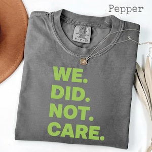 Puede incluir: Camiseta gris color pimienta con el texto "WE. DID. NOT. CARE." en verde brillante. La camiseta tiene cuello redondo y mangas cortas. Un collar plateado con un pequeño colgante está sobre la camiseta.