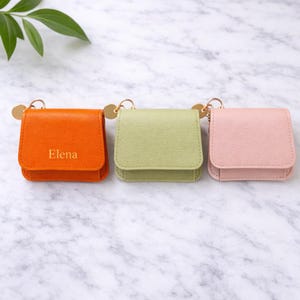 Könnte beinhalten: Drei kleine, quadratische Etuis in Orange, Grün und Rosa. Jedes Etui hat einen goldfarbenen Schlüsselanhänger und einen kleinen Klappverschluss. Auf dem orangefarbenen Etui ist der Name "Elena" in Gold aufgedruckt.