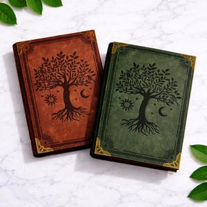Può includere: Due diari rilegati in pelle, uno marrone e uno verde. Entrambi presentano un design ad albero in rilievo con dettagli di sole, luna e stelle. Gli angoli dorati aggiungono un tocco decorativo. I diari sono su uno sfondo di marmo.