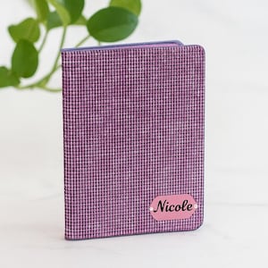 Peut inclure: Un portefeuille violet avec un motif texturé en forme de grille. Le portefeuille comporte une petite étiquette ovale rose avec le nom "Nicole" en écriture cursive. Le portefeuille est debout, mettant en valeur son design.
