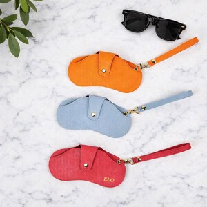 May include: Three coloured eyeglass cases in orange, blue, and red, each with a matching wrist strap and gold-tone hardware. The red case has the initials "ELO" printed on it. Black sunglasses are at the top.