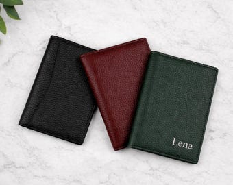 May include: Three leather passport holders in black, burgundy, and green. The green holder has the name "Lena" printed on it in white. The holders are arranged on a white marble surface with green leaves in the background.