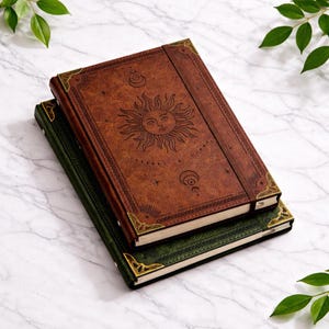 Pode incluir: Dois cadernos empilhados com desenhos celestiais. O caderno superior é marrom com um desenho de sol e estrela, e o caderno inferior é verde. Ambos têm cantos de latão e um fecho de faixa elástica.